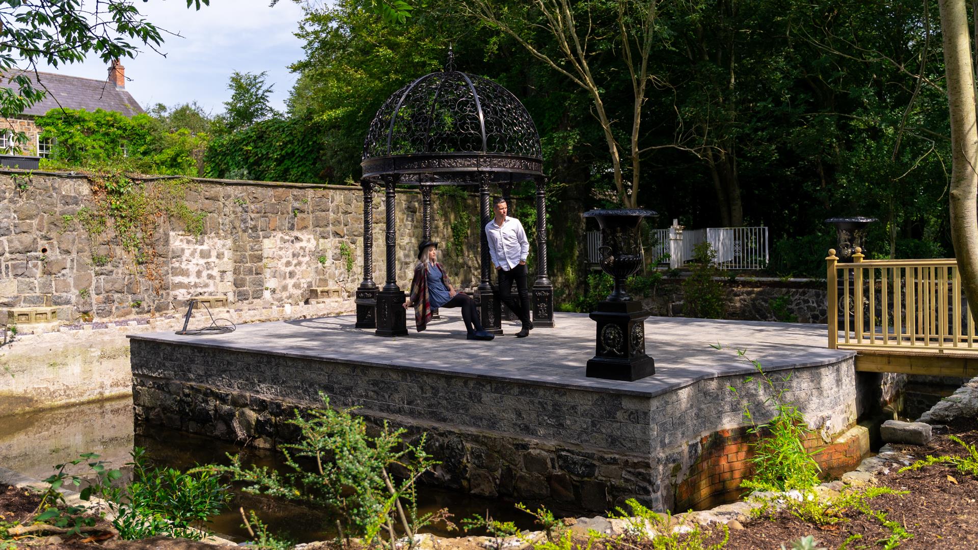 A man and woman resting under a canopy on a small manmade island in a garden.