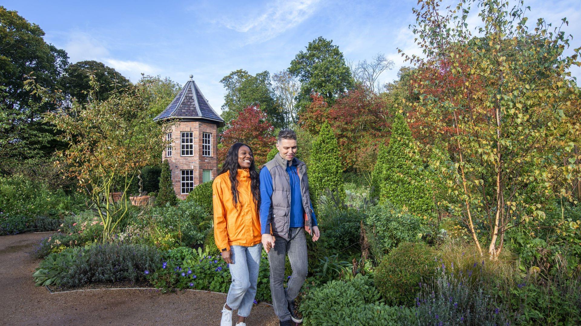 A couple walking on a garden path with lots of trees and other plants.