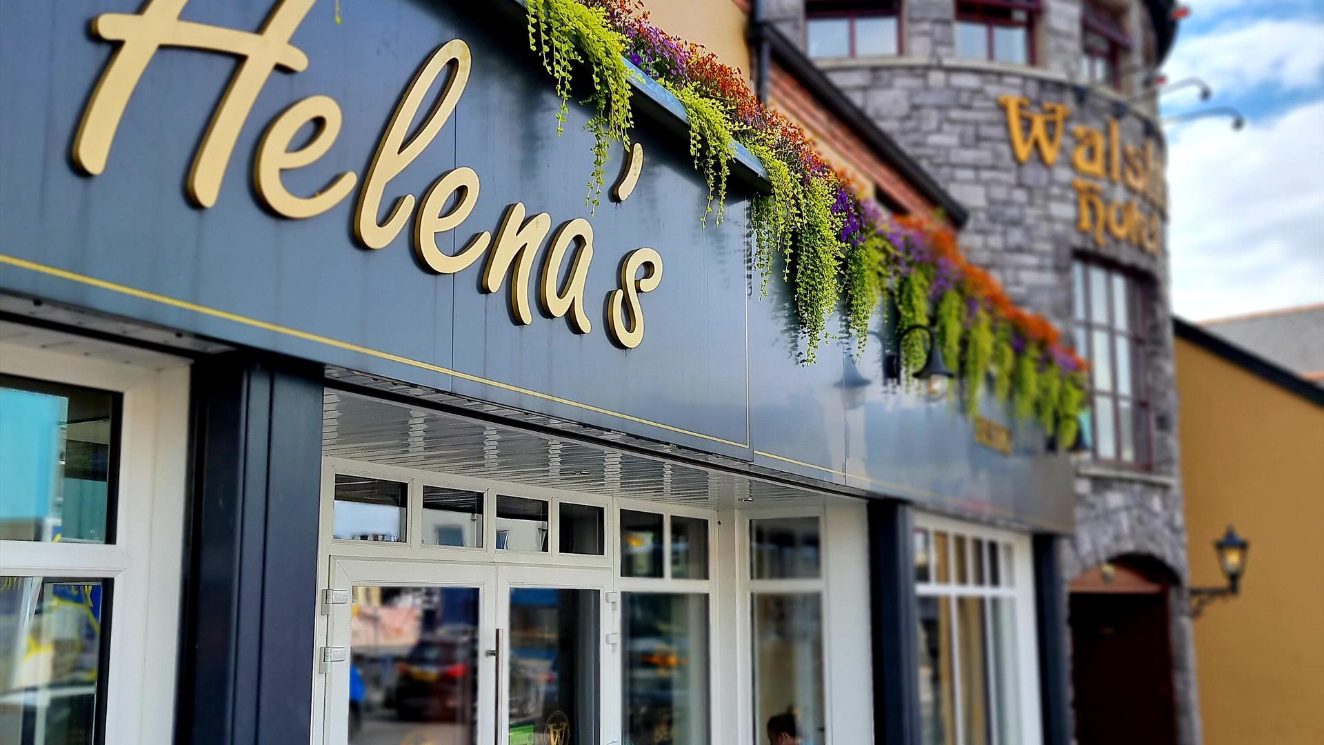 Exterior of Helena's Coffee Shop and Bistro, featuring a modern black and white facade with gold lettering. Colorful hanging plants decorate the top o