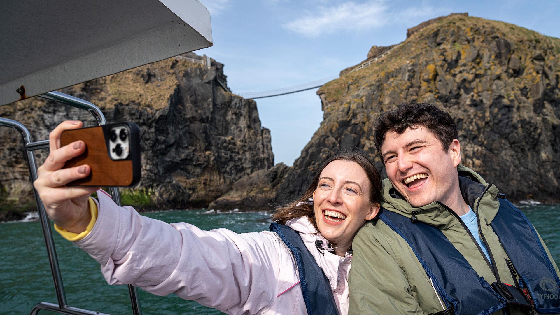 Couple on a boat taking a selfie and enjoying the Giant Shipwrecks of the North Coast experience with Carrick-A-Rede Rope Bridge in the background