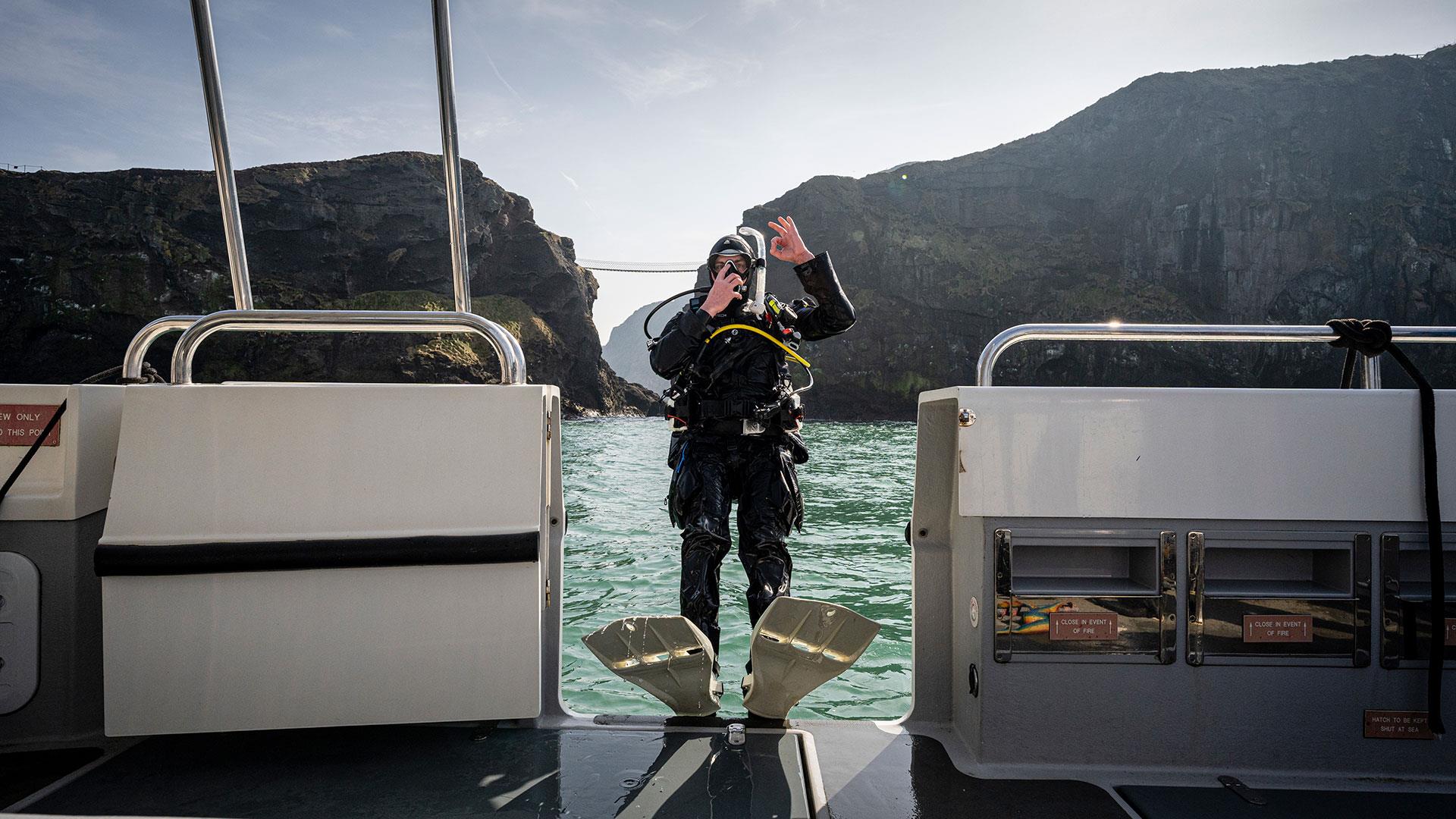 Girl on a boat in scruba diving gear falling backwards with Carrick-a-Rede Rope Bridge in the background as part of a scuba diving experience with Aqu