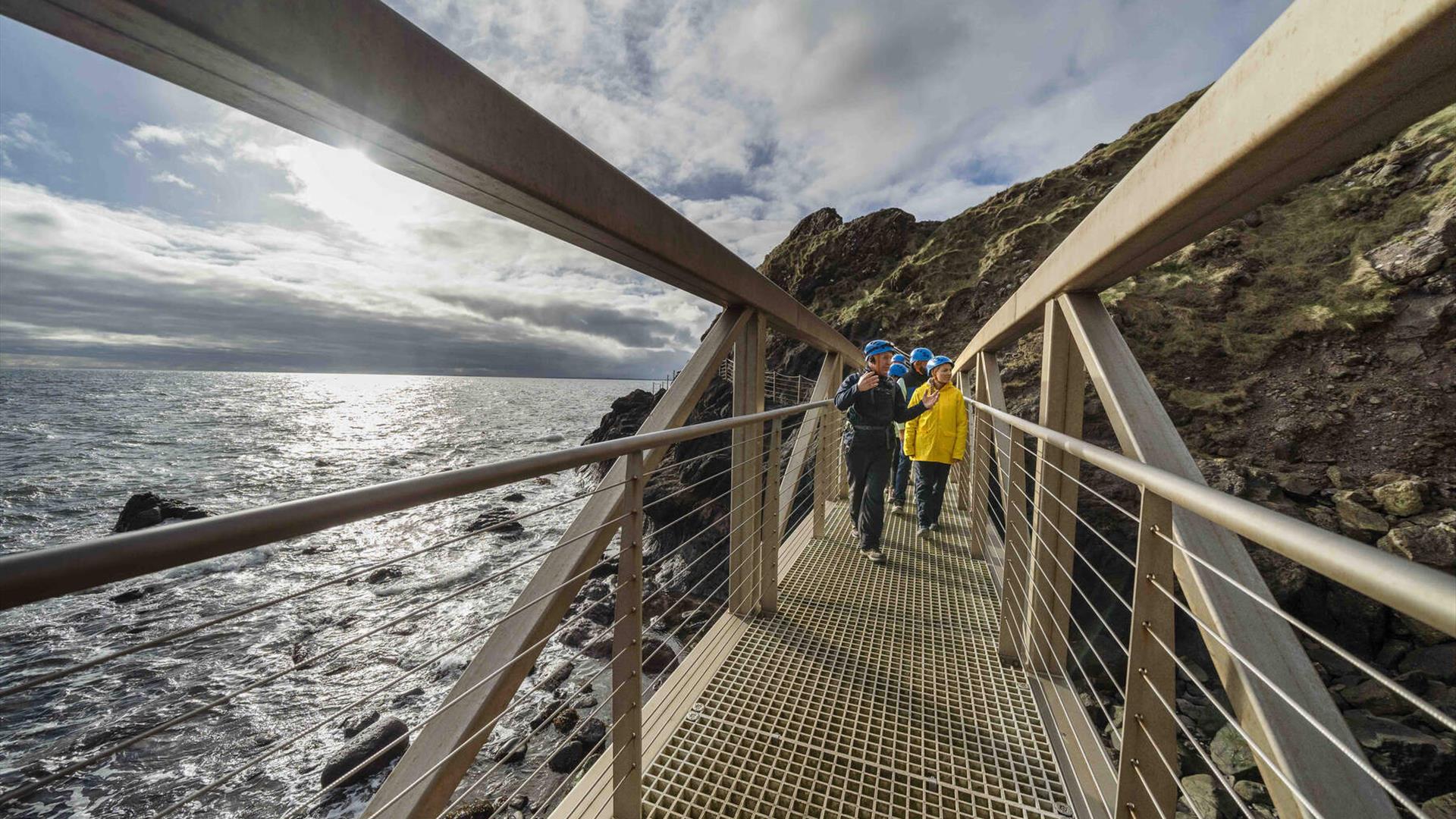 Sun shines on group as they walk along metal bridge at The Gobbins