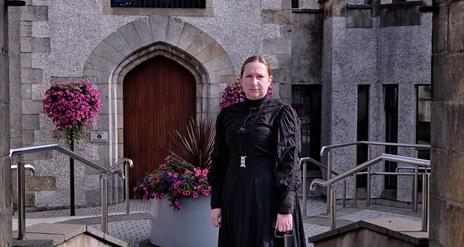 Image of woman standing under the stone arch of the Bridewell, Magherafelt.  She is dressed in a long black dress from 1800's and holds a large brass