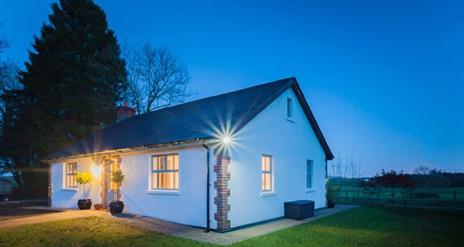 A cottage at night beside a country feild in front of a large tree.