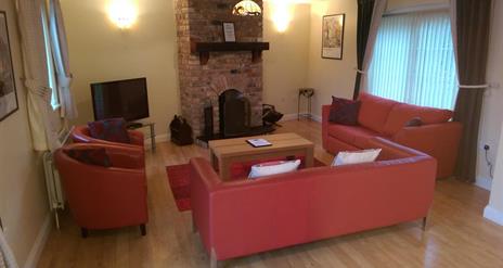 Image shows lounge area with sofas, chairs, TV and fireplace. Two windows in the room.