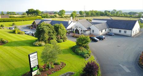 Image shows hotel and grounds with grass area and sign re hotel name. Also shows parking areas