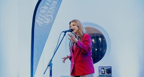 a woman sings in to a microphone on stage with a branded Atlantic Sessions flag beside her