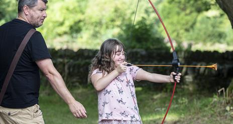 Girl shooting bow and arrow with her father behind her.