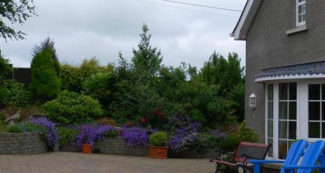 A paved area with garden furniture, trees and purple flower bushes.