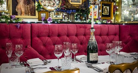 A table with red felt booth seating set with wine glasses and a champagne bottle holding a candle.
