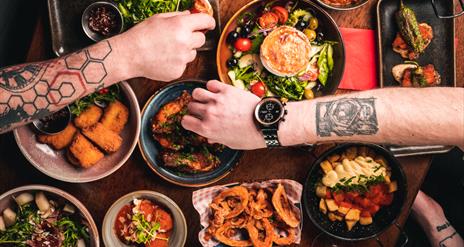 People with tattooed arms reaching over a table for tapas food.