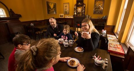 Family of 3 children and 2 adults sitting eating and drinking tea around a wooden dining table in the railway musuem cafe