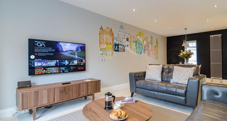 A living room with grey leather sofas, a coffee table with coffee and croissants and a TV with Netflix.