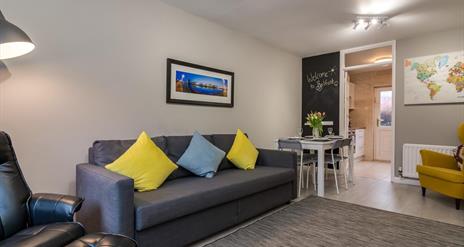 A photo of a living room with a sofa and chair which leads to a dining table and kitchen entrance.