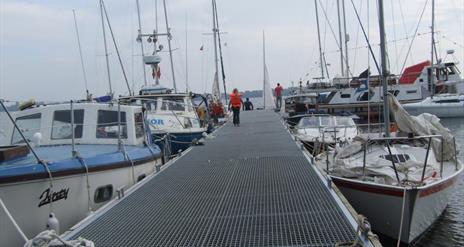 Image shows a walkway and various types of boats in a marina