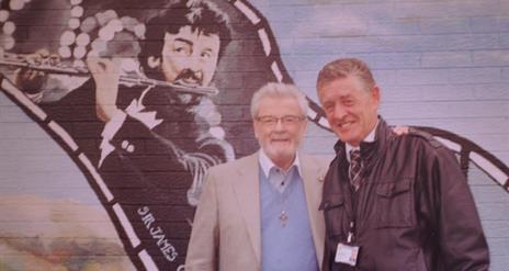 Two men standing in front of a mural of Sir James Galway.