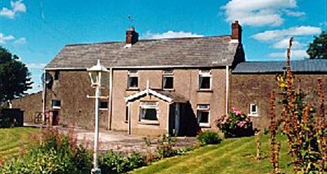 A large farmhouse with a front garden filled with flowers and a lamp.