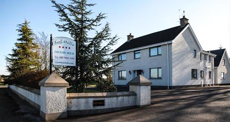 A roadside view of a large country house with a sign and trees outside.