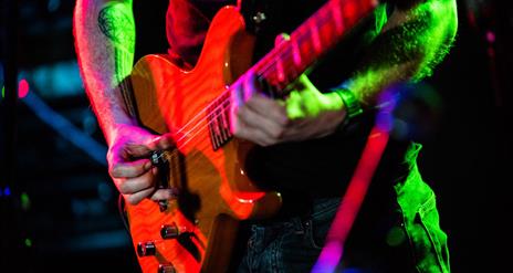 Close up image of an electric guitar being played. The guitarist has a tattoo on his arms and is wearing dark jean and a dark coloured t shirt.