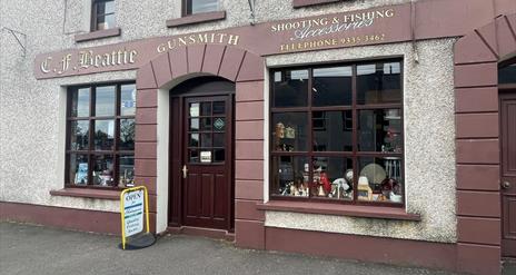 Brown and cream exterior of CF Beattie with sign stating Shooting & Fishing Accessories / Gunsmith plus antique displays in windows