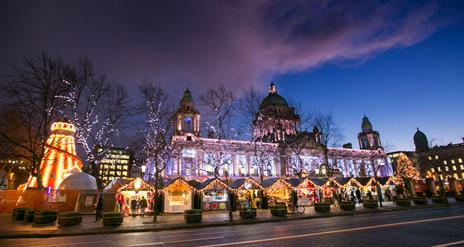 Belfast City Hall light up at night with Christmas market stalls in foreground in front of the City hall front gates.