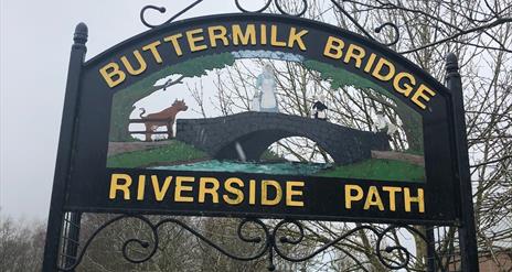 Black metal Buttermilk Bridge Riverside Path sign with writing in yellow and image of a bridge with cow and milk maid crossing