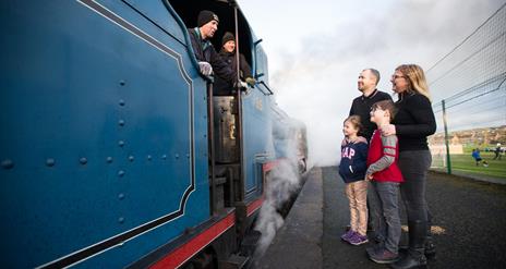 A family of four standing on the platform talking to the driver of a blue steam train