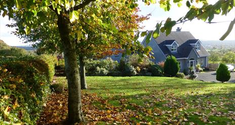 A lawn with falling leaves of a large grey house in the hills.