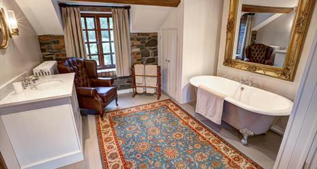 Image shows large bathroom with roll top bath and large mirror above it. Large rug. armchair, wash basin with storage underneath