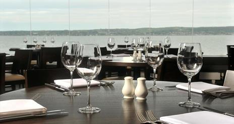 Waterfront restaurant with many tables set with wine glasses, cutlery and salt and pepper shakers.