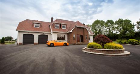 Image shows front of property with orange coloured super car in the large driveway