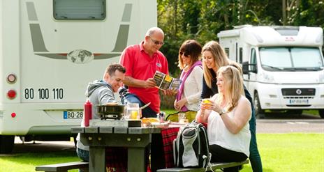 Image shows family and friends sitting at picnic table with caravan in the background