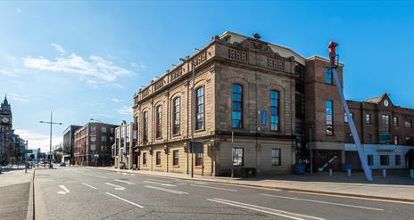 The Corn Exchange, a large light-coloured stone building with arched windows, stands on a quiet city street under a clear blue sky. There are addition