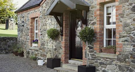 Image shows outside of stone property with bay tree plants at either side of front door