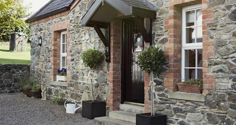 Image shows outside of stone property with bay tree plants at either side of front door