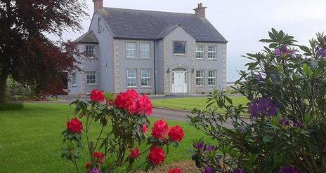 The front of a large grey house behind some colourful flowers.