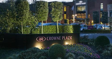 Image of the outside of the Crowne Plaza Belfast hotel. Flower bed with "Crowne Plaza" sign in the foreground, hotel and parking lot in the background