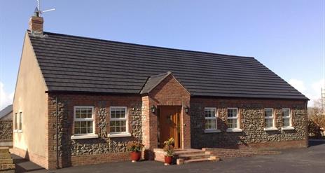 Image shows front of detached cottage which has 6 windows and front door with a few low steps