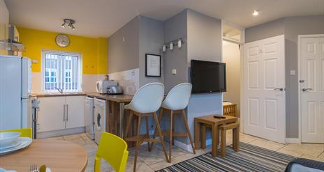 A photo of a kitchen which wraps around a wall with a tv to a living space.