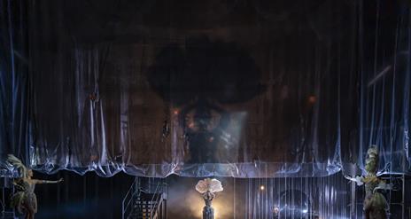 A transparent curtain rises on a dark set, with a smoky spotlight highlighting the shadow of a showgirl with a huge feather headdress walking centrall