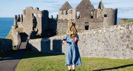 Person in a dress wearing a traditional 'mummers' mask plays the violin outside Dunluce Castle