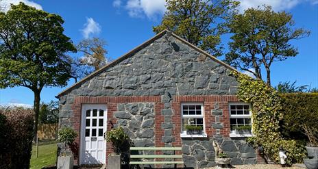 Image shows stone cottage covered in ivy at the right hand side plus white front door and bench outside