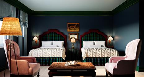 Spacious hotel room with two double beds featuring tartan headboards, soft cream bedding, and classic wood furniture. The room includes pink armchairs