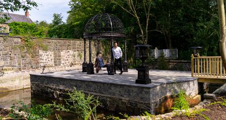 A man and woman resting under a canopy on a small manmade island in a garden.