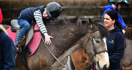 a child on horseback leans forward to hug a horse, while an instructor watches on