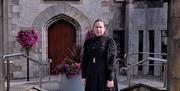 Image of woman standing under the stone arch of the Bridewell, Magherafelt.  She is dressed in a long black dress from 1800's and holds a large brass