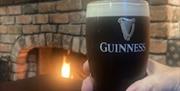 A pint of Guinness rests on a bar table with a hand grasping it. In the background , an open fire burns in a large brick fireplace.