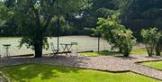 A lawn surrounded by gravel paths with trees and a garden table leading to a tennis court.