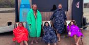 a group of five people stand and relax on deck chairs in front of the mobile sauna on the beach