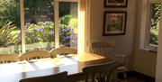 A wooden dining room table with chairs in a room with windows viewing a flowery garden.